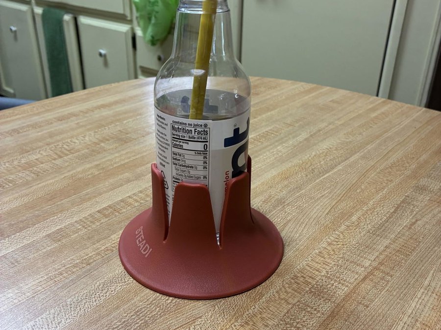 A clear glass drink bottle with a yellow straw sitting in a brick-red drink base on a light oak kitchen table, pale green cabinets and a kitchen counter behind